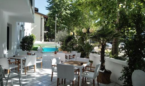Terrazza con tavoli bianchi, piscina e alberi verdi.